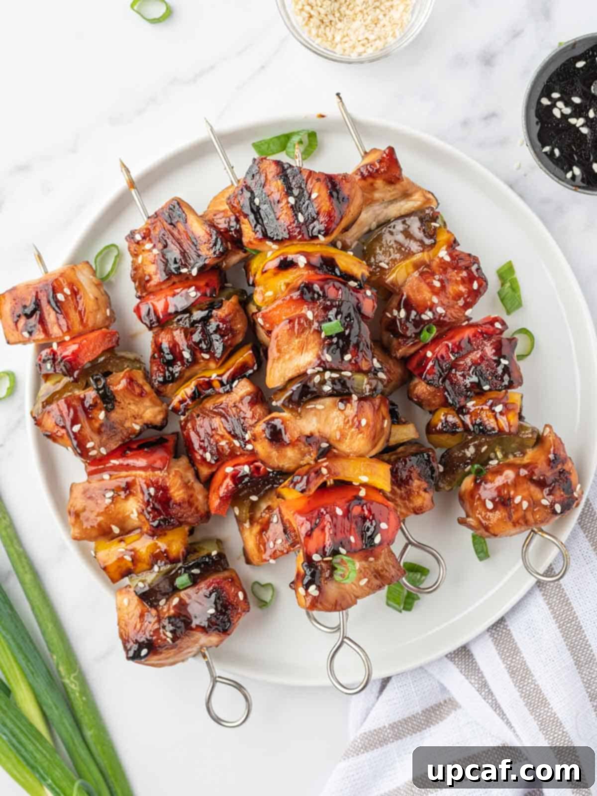 Sweet and Sticky Teriyaki Chicken Skewers 2 A pile of grilled chicken teriyaki skewers on a round platter, garnished with sesame seeds and fresh herbs.