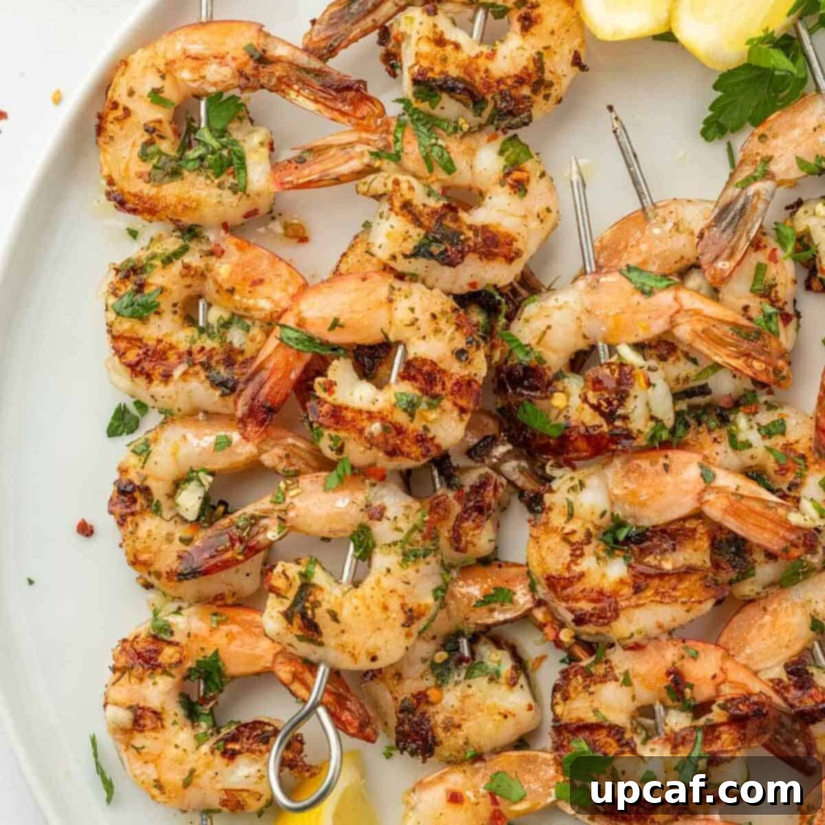 Flame-Kissed Garlic Prawns 7 Shrimp kabobs with garlic arranged on a white platter with lemon wedges.
