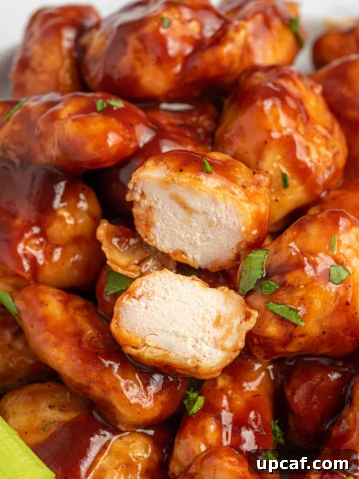 A close-up shot of a baked BBQ chicken bite sliced in half, resting on a pile of other sauced chicken, revealing the tender interior.