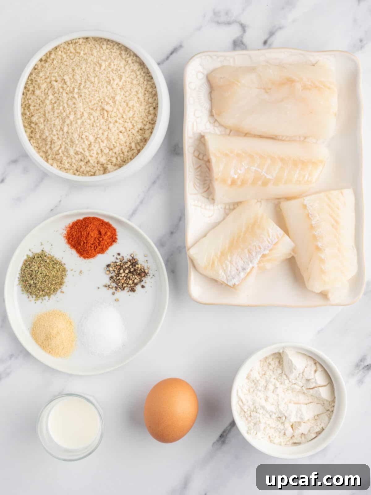 All ingredients for crispy air fryer cod laid out on a cutting board, including cod fillets, flour, eggs, panko, and spices.
