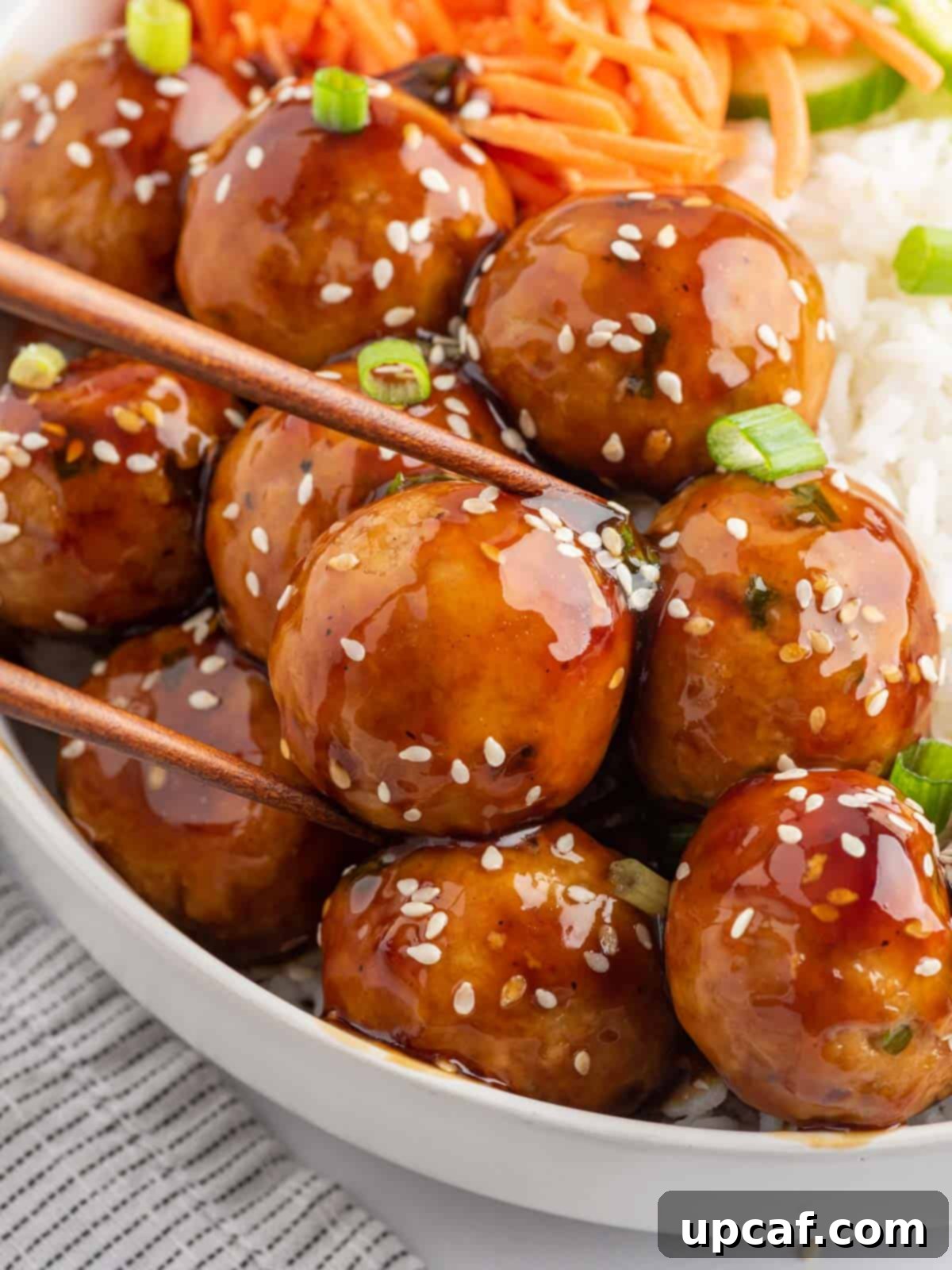 Chopsticks picking up a single teriyaki-coated meatball from a plate.