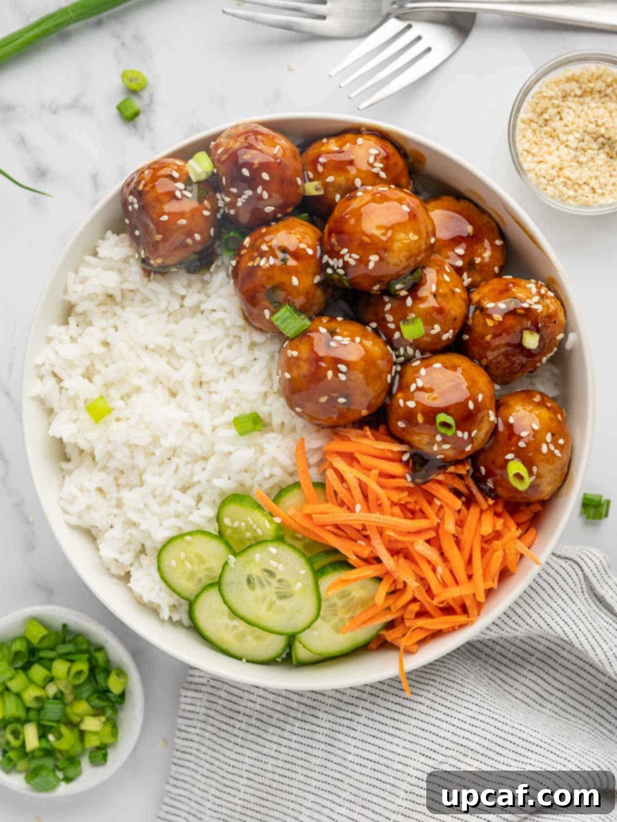 Teriyaki meatballs on a plate with white rice, sliced cucumber, and julienned carrots, ready to be served.