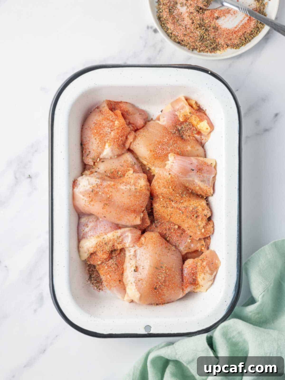 Seasoning raw chicken thighs in a dish with a mix of herbs and spices.
