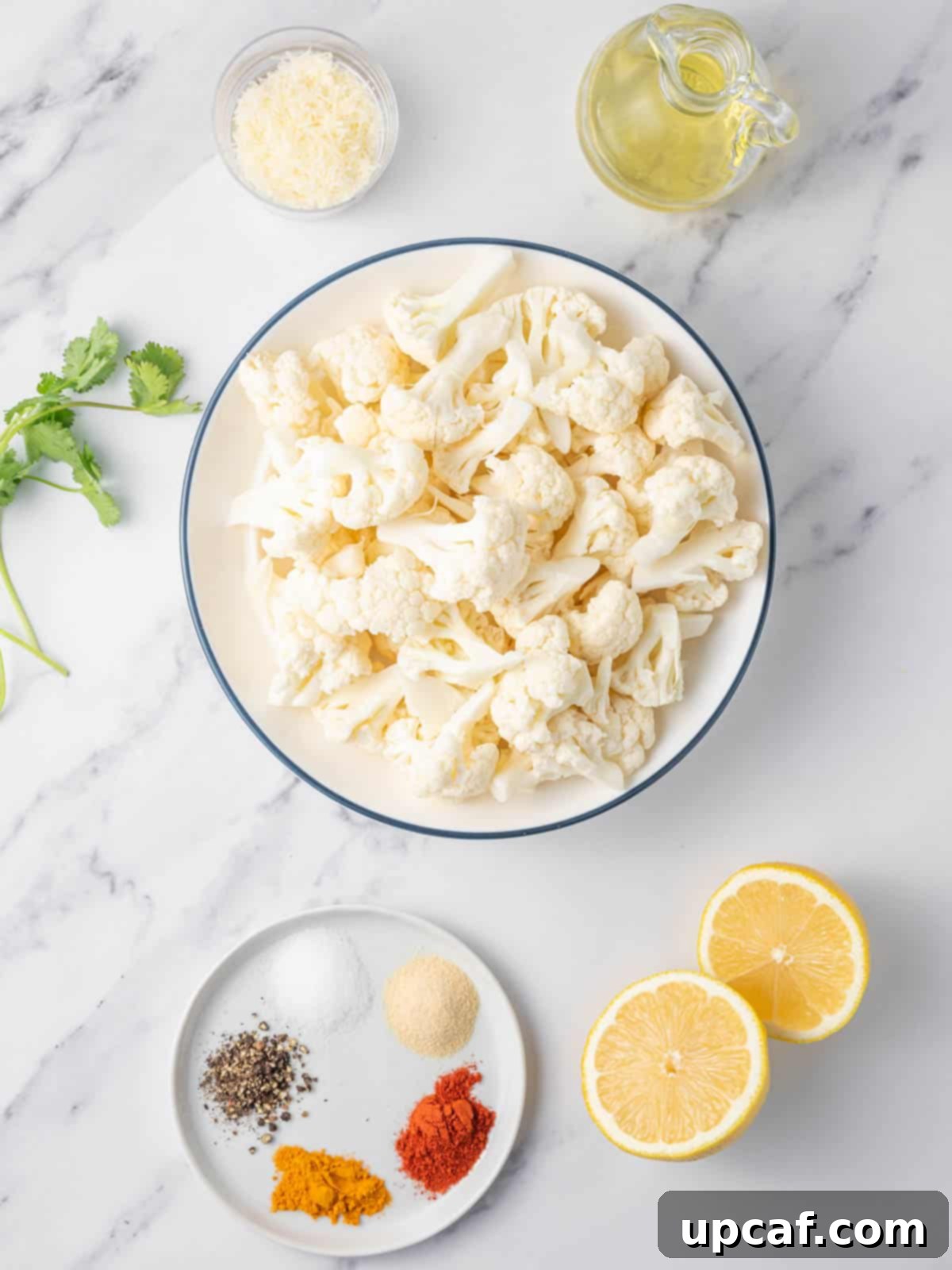 A selection of fresh ingredients laid out, including a head of cauliflower, various spices, olive oil, parmesan cheese, and cilantro, ready for air frying.