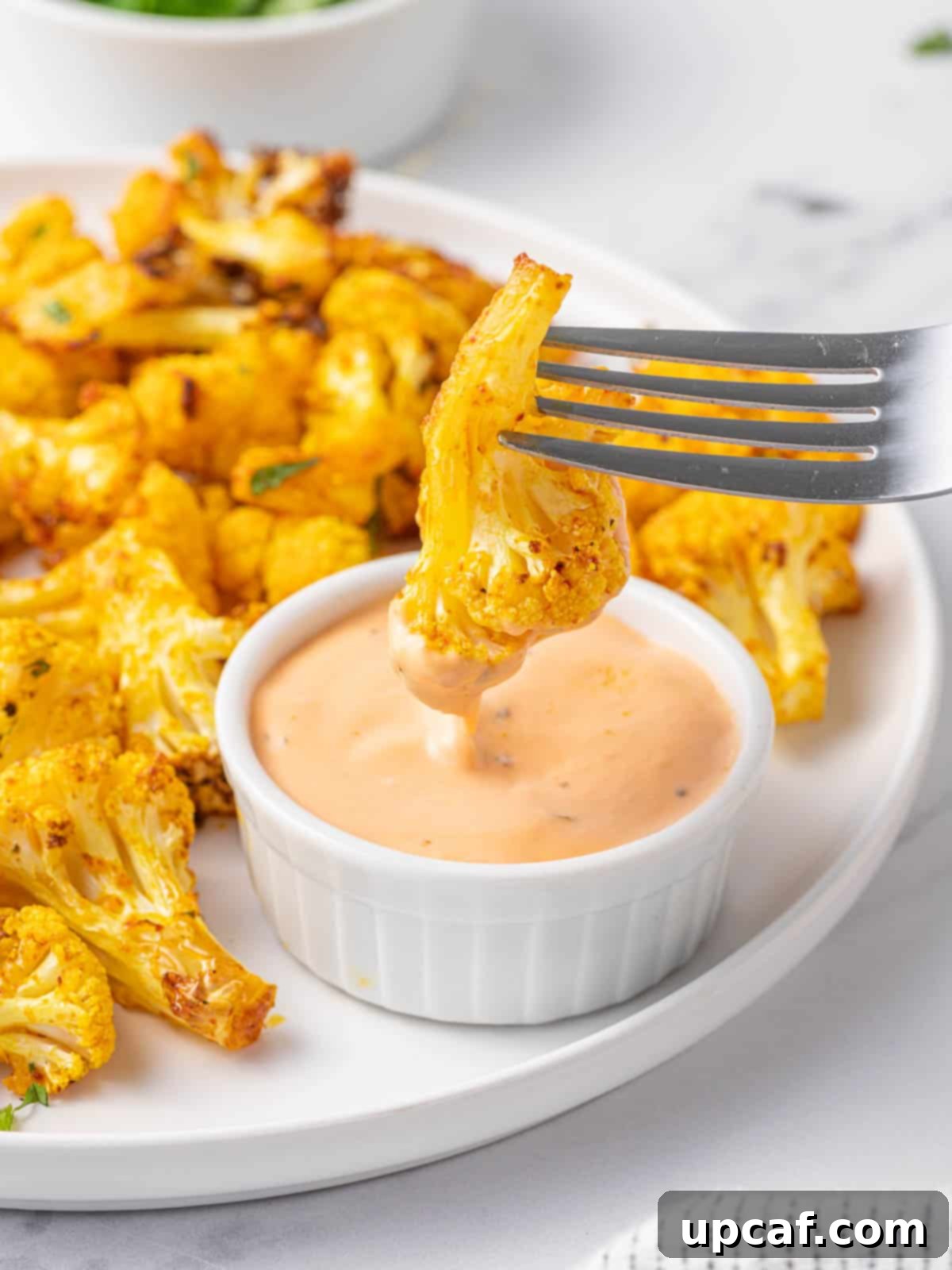 A fork piercing an air fryer cauliflower bite, poised above a dipping sauce, highlighting the interactive enjoyment.