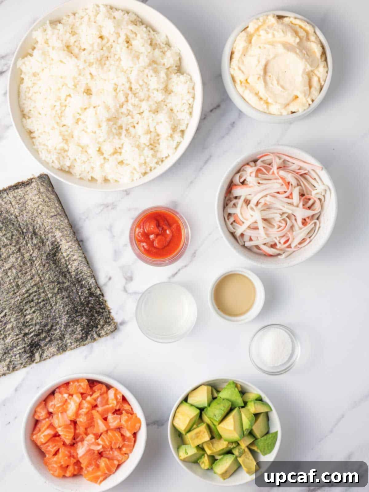 A selection of sushi burger ingredients laid out on a kitchen counter: cooked sushi rice, fresh salmon, imitation crab, avocado, nori sheets, sriracha, mayonnaise, lime juice, and green onions.