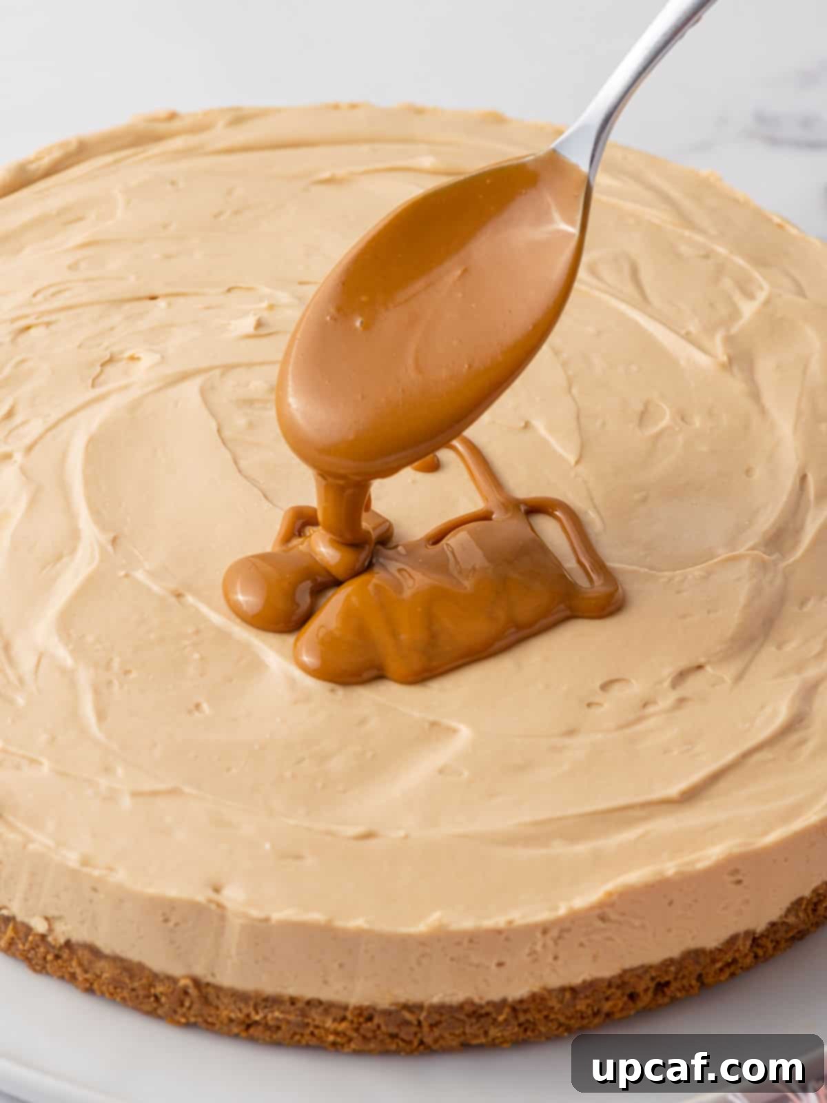 A spoon drizzles melted biscoff spread onto a cheesecake, illustrating the topping step.