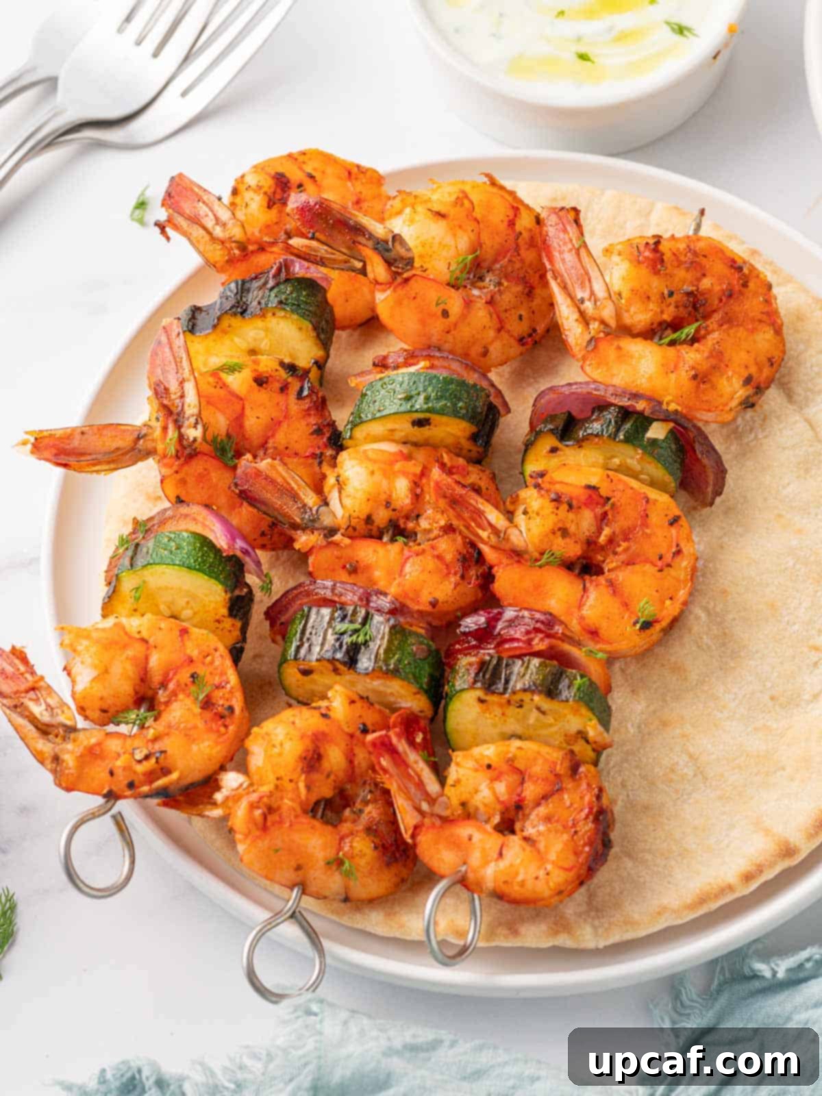 Golden Oven Shrimp Skewers 8 Three shrimp skewers served on a piece of pita bread with a side of dipping sauce and a lemon wedge.