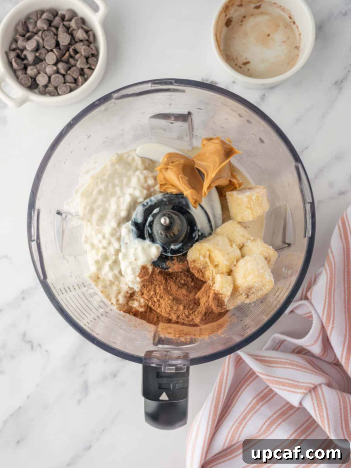 Ingredients for chocolate peanut butter cottage cheese ice cream, including cocoa powder, peanut butter, frozen banana, and cottage cheese, being blended in a food processor.