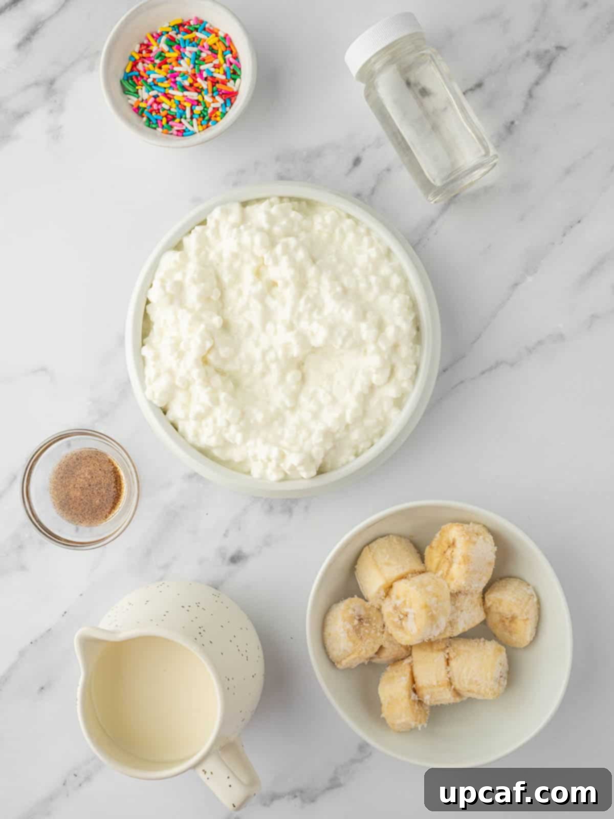 A selection of ingredients laid out for vanilla cottage cheese ice cream, including cottage cheese, heavy cream, vanilla syrup, vanilla paste, and frozen banana slices.