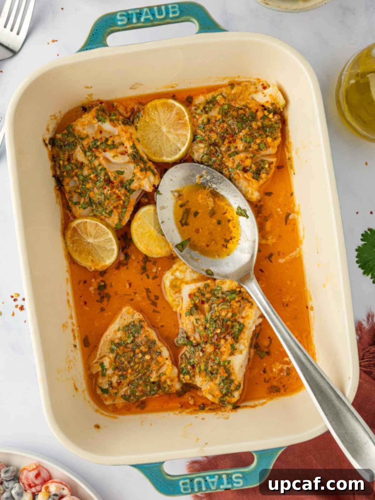 A spoon ladles rich buttery herb lime cod marinade over cooked fish in a casserole dish, highlighting the glistening finish.