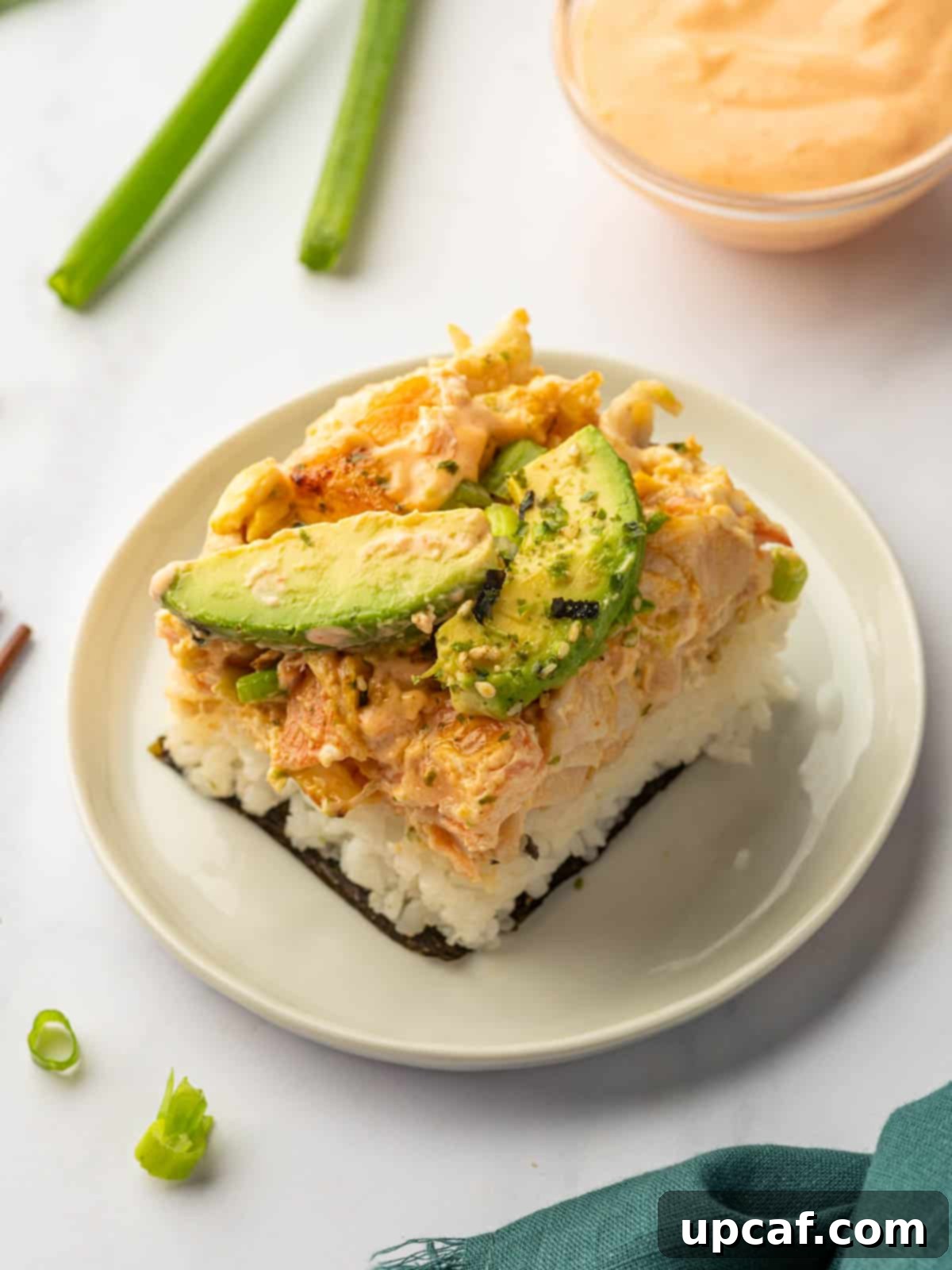A square piece of baked salmon sushi on a white plate, garnished with avocado, green onions, and a drizzle of spicy mayo, with a nori sheet on the side.