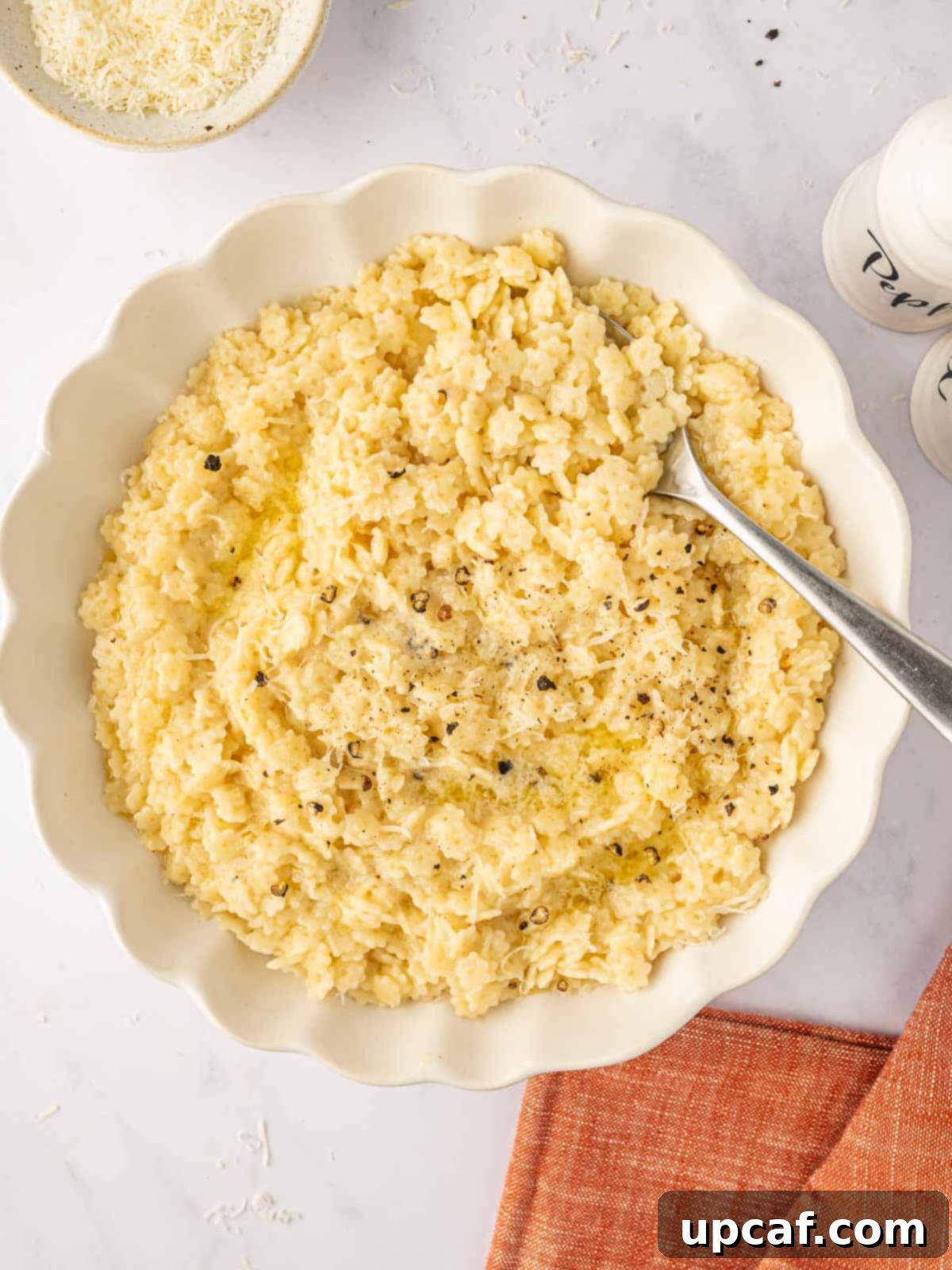 A spoon rests in a white bowl of pastina pasta, garnished with fresh herbs and grated Parmesan cheese.