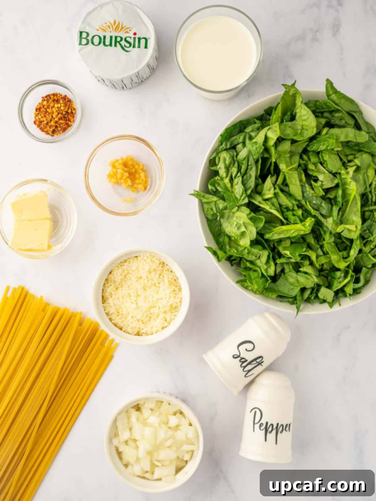 A flat lay photograph showing all the fresh ingredients needed for Boursin cheese pasta: linguine, Boursin cheese, heavy cream, spinach, garlic, onion, butter, and seasonings.