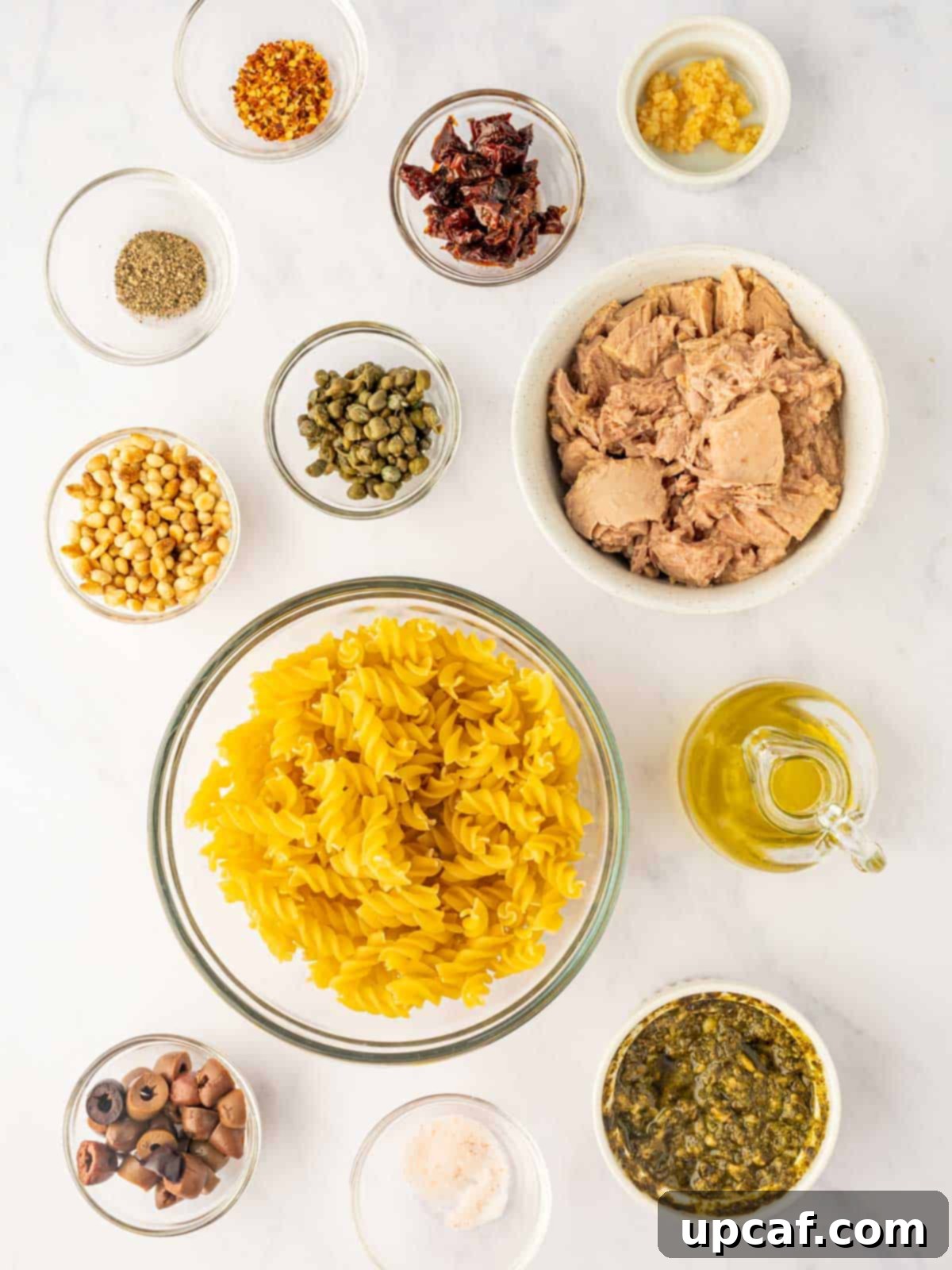 Mediterranean Tuna Pesto Pasta 3 Assorted ingredients for tuna pesto pasta laid out on a white surface, including fusilli pasta, a can of tuna, a jar of pesto, sun-dried tomatoes, capers, garlic, and olives.