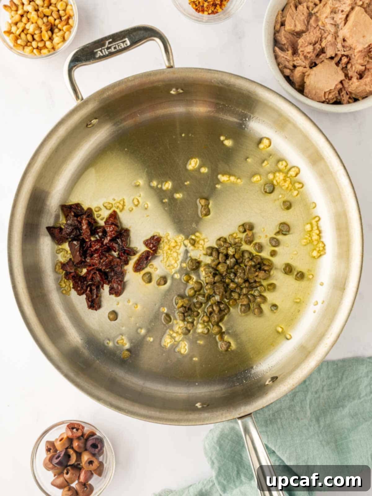 Mediterranean Tuna Pesto Pasta 4 Minced garlic, chopped sun-dried tomatoes, and capers being sautéed in a large skillet with olive oil over medium heat.