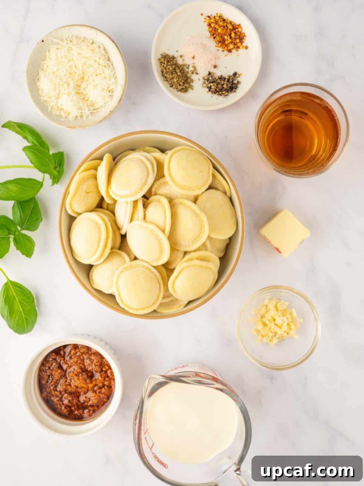 A vibrant selection of ingredients for cheese ravioli pasta, including fresh basil, sun-dried tomato pesto, cream, and cheese, artfully arranged on a counter.