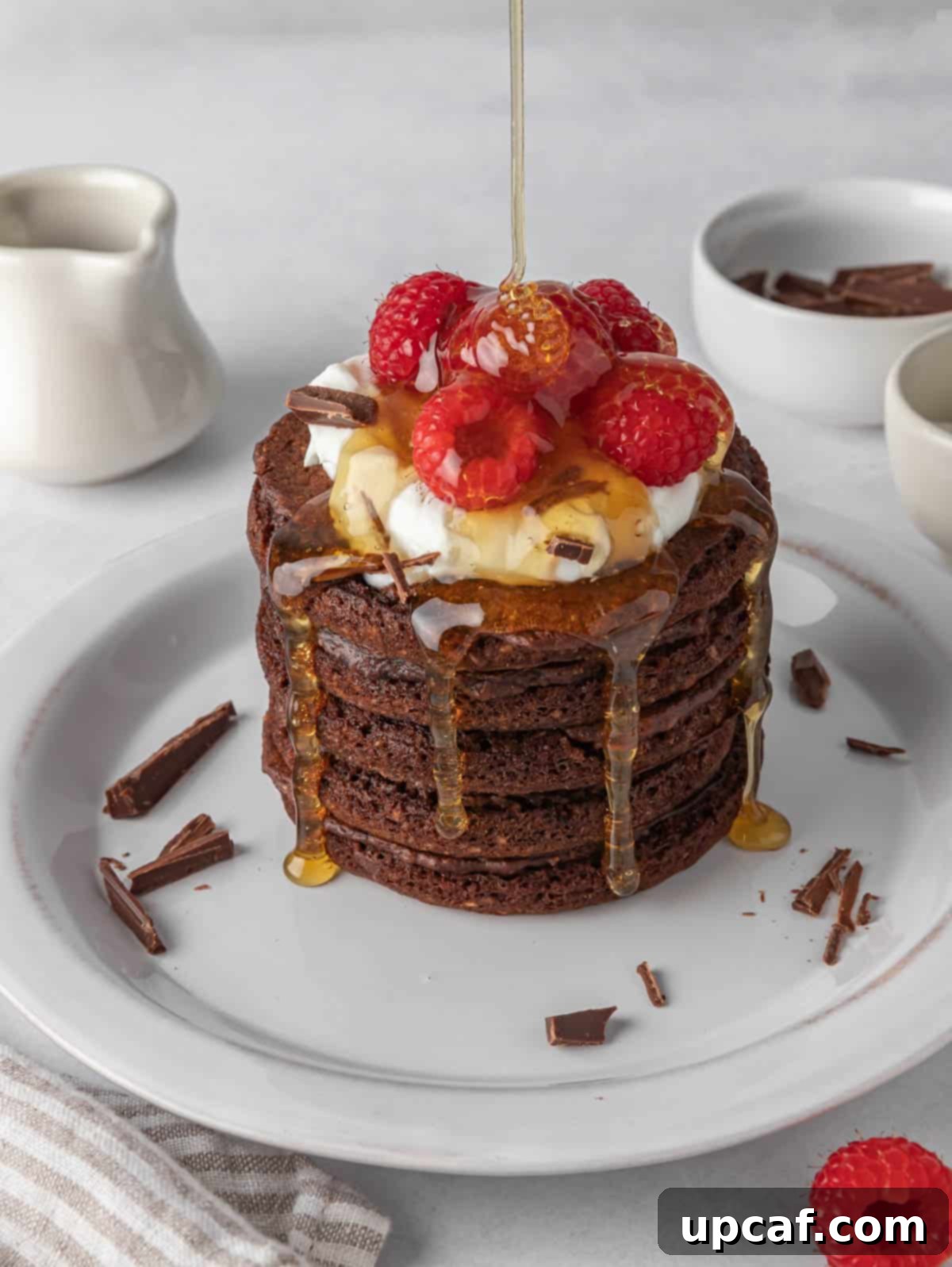 Ultimate Chocolate Protein Stacks 10 Pancakes on a white plate with syrup being drizzled over them.