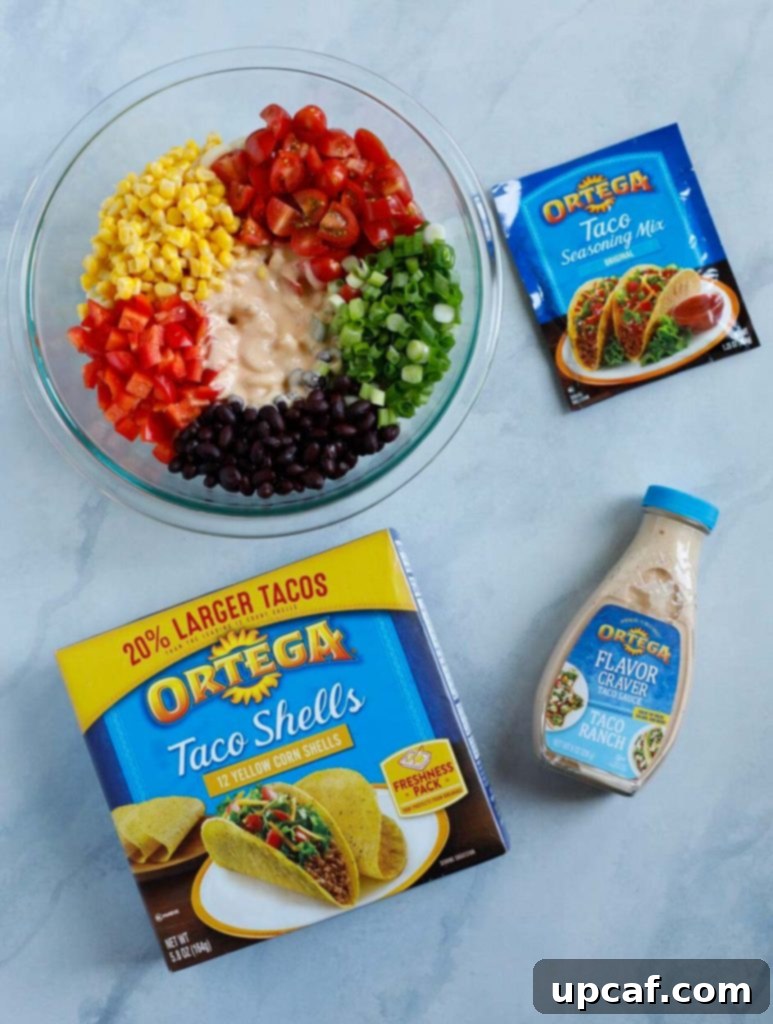 Array of fresh ingredients laid out for Taco Ranch Pasta Salad, including elbow pasta, grape tomatoes, corn, black beans, green onions, red bell pepper, lime, salt, Ortega taco seasoning, Ortega Taco Ranch Sauce, shredded cheese, and avocados.