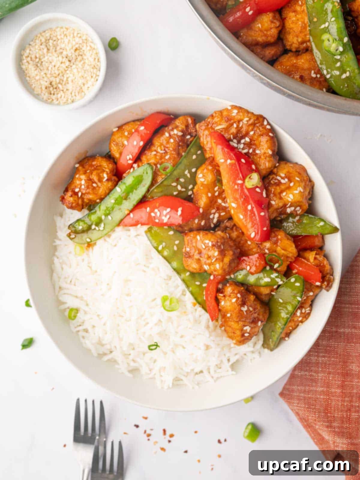 A plate with a generous serving of crispy honey chili chicken, rice, and fresh vegetables, garnished with sesame seeds and green onions.
