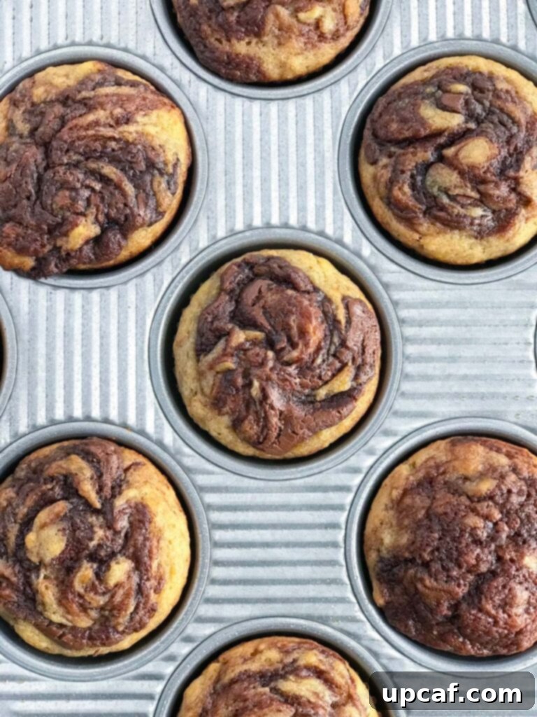 Nutella Banana Swirl Muffins in Pan A fresh batch of golden brown Nutella Swirl Banana Muffins in a muffin pan, cooling down.