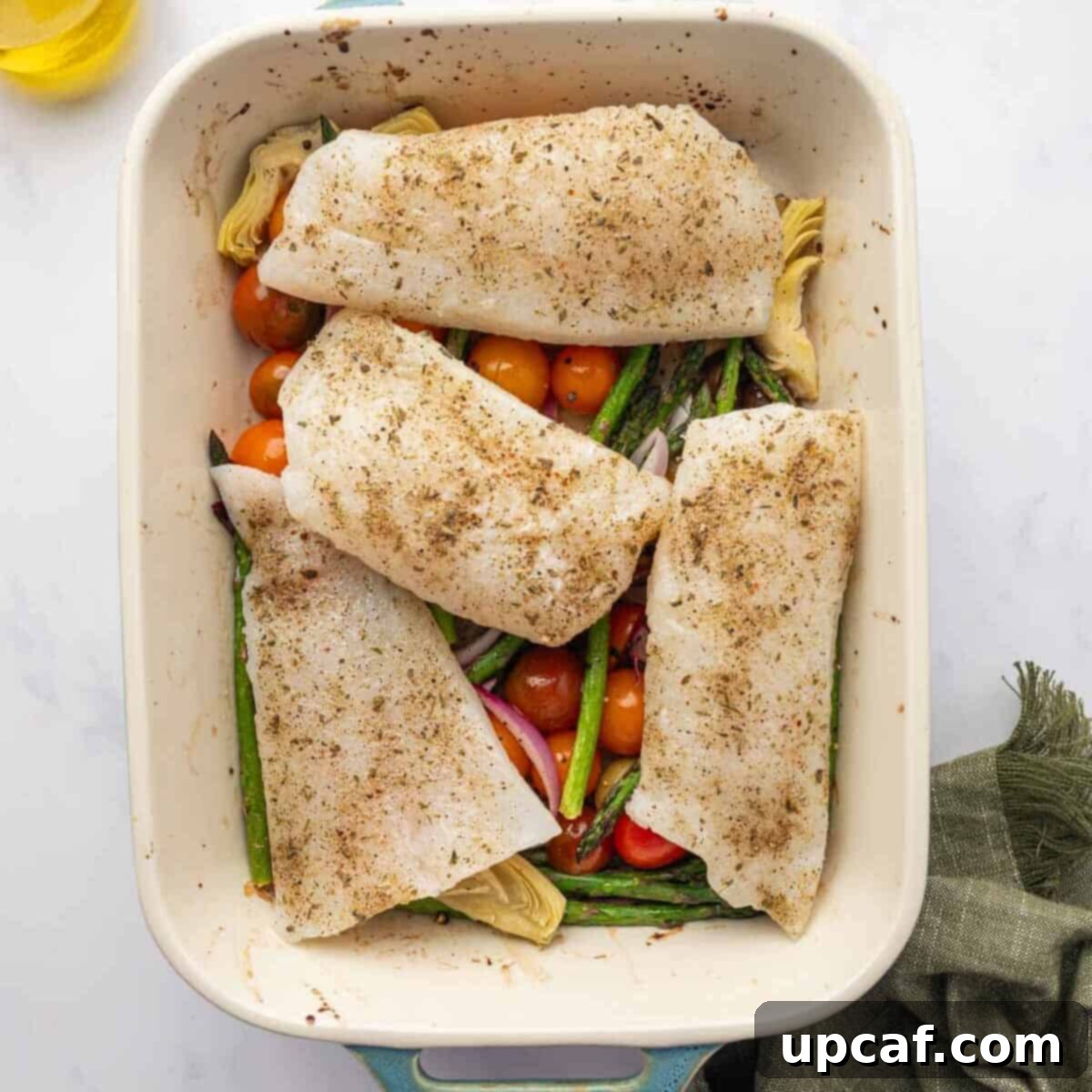Seasoned cod is placed on top of vegetables in a baking dish.