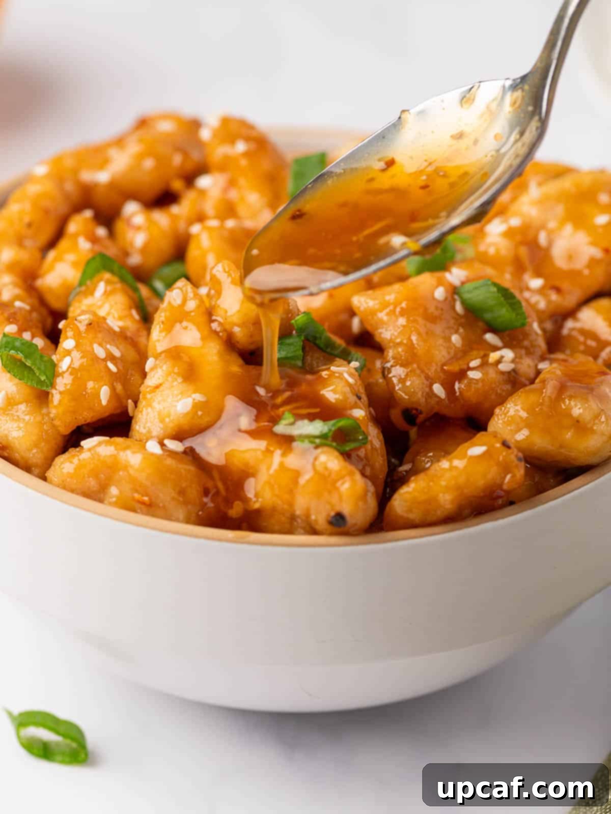A spoon drizzling homemade orange chicken sauce onto a bowl of crispy fried chicken pieces.
