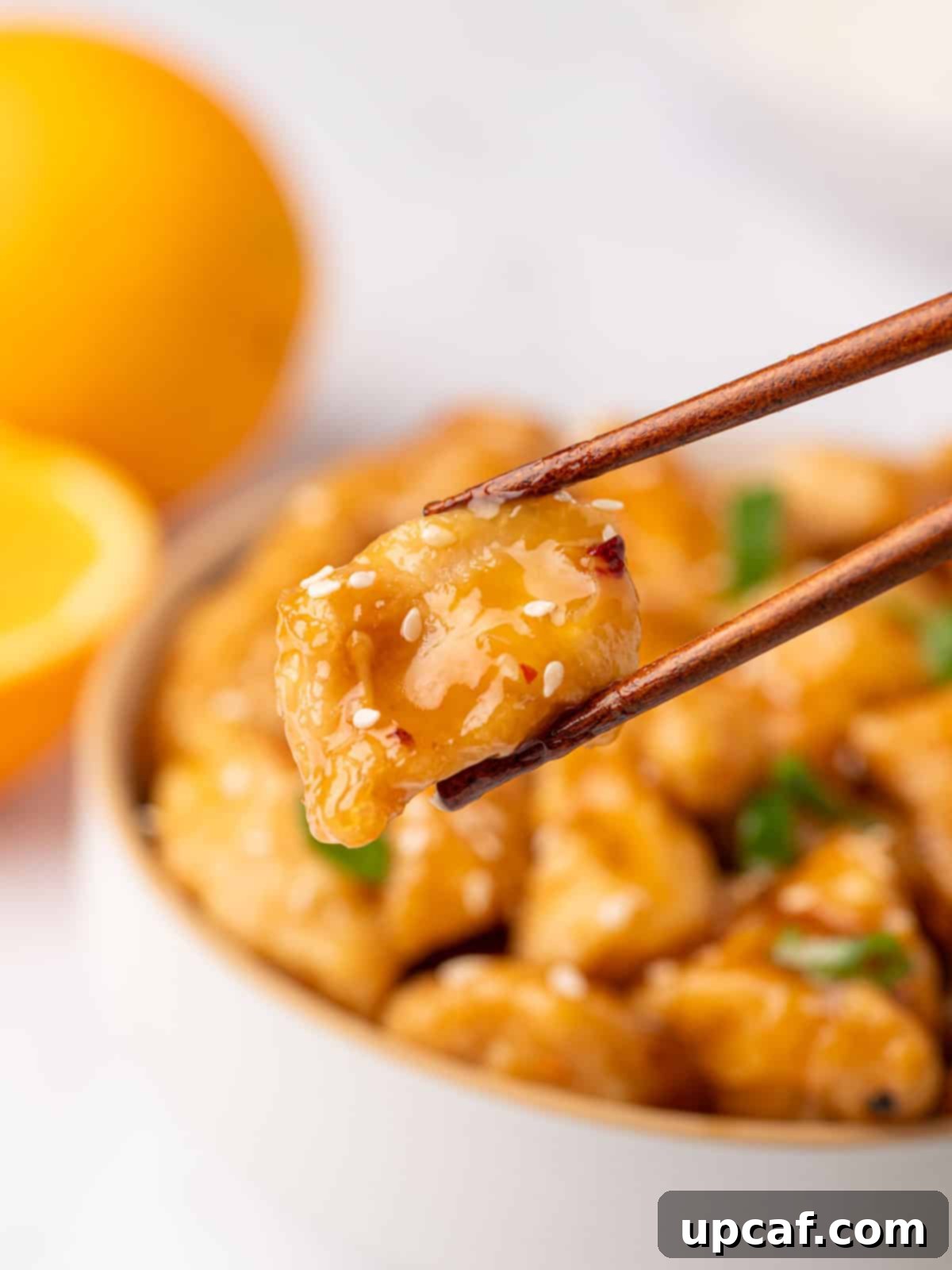 Chopsticks picking up a piece of fried chicken coated in homemade orange sauce from a bowl.