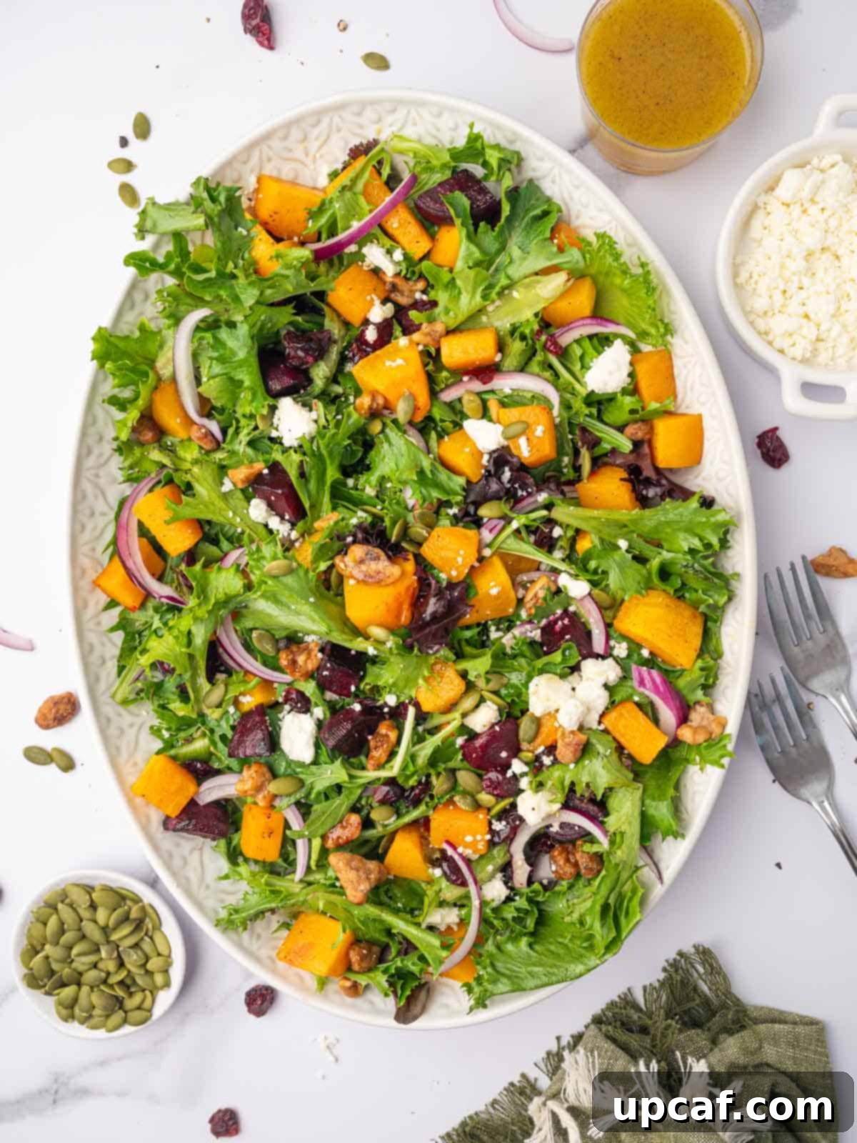 Roasted Butternut Feta Salad 2 A large platter with a green salad topped with golden roasted butternut squash, thinly sliced red onion, and crumbled feta cheese, garnished with fresh herbs and colorful seeds.