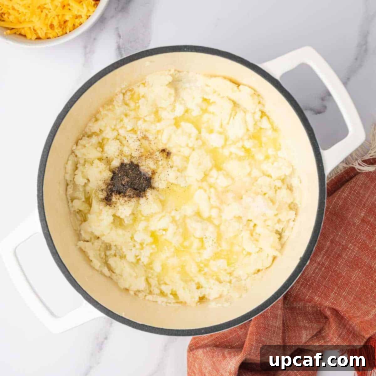 Decadent Cheesy Mash 6 Seasoning is added to mashed potatoes in a pot, with butter and half-and-half nearby.