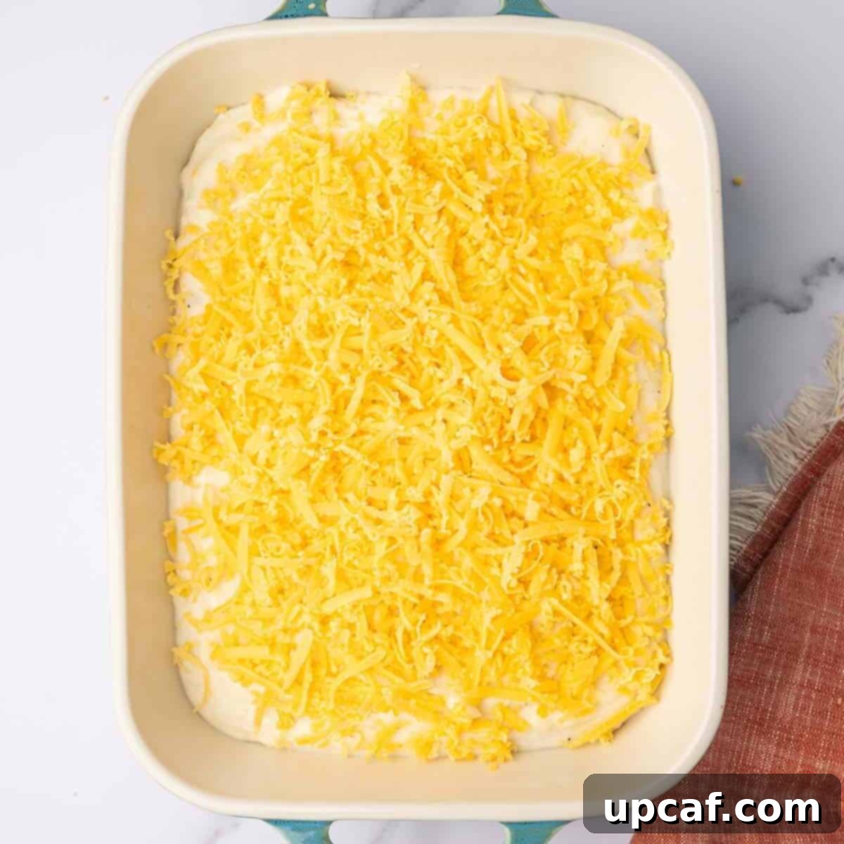Decadent Cheesy Mash 9 Additional shredded cheese is sprinkled on top of mashed potatoes in a baking dish before baking.