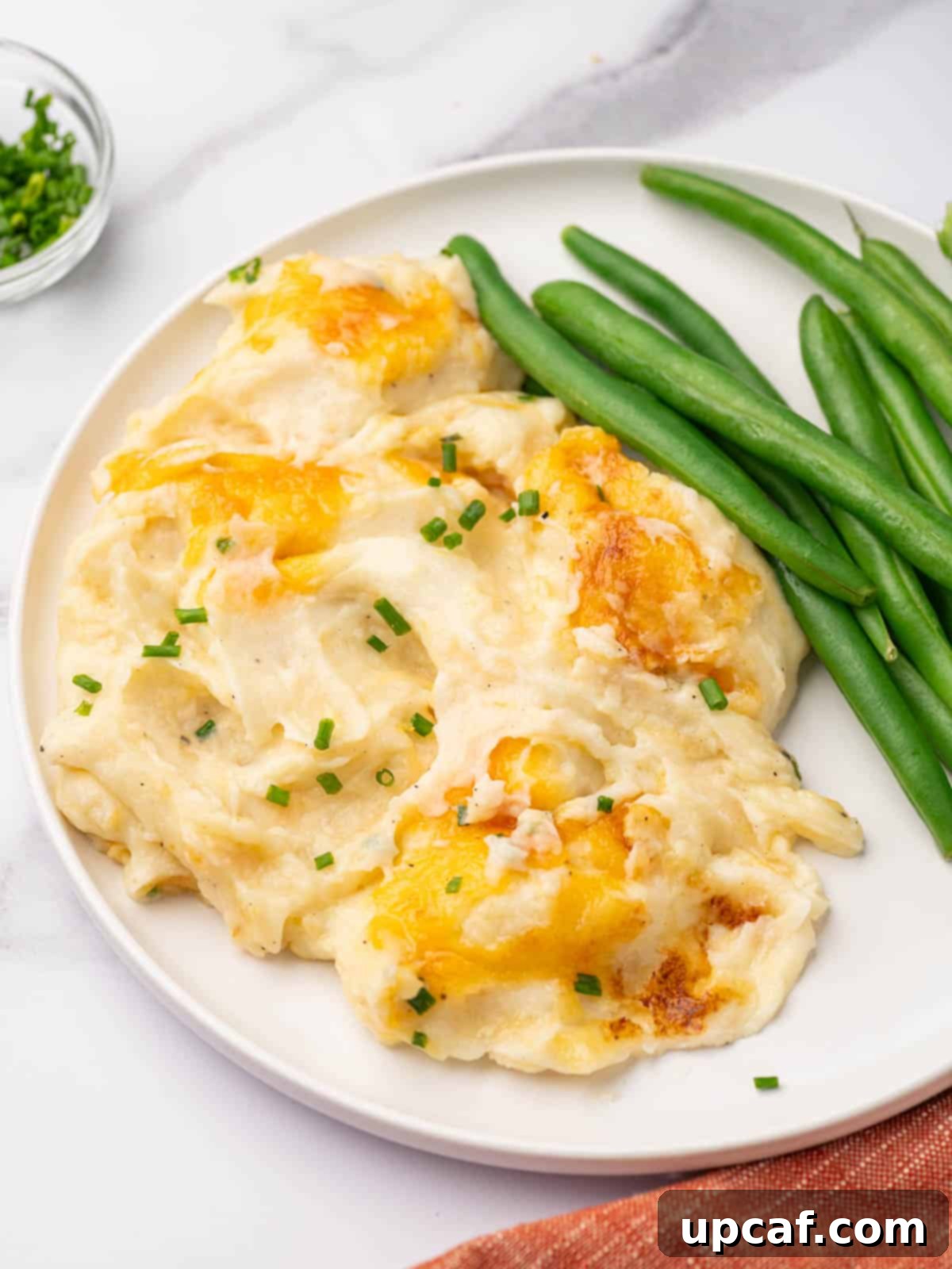 Decadent Cheesy Mash 10 Cheesy mashed potatoes on a white plate with a side of fresh green beans, garnished with chives.