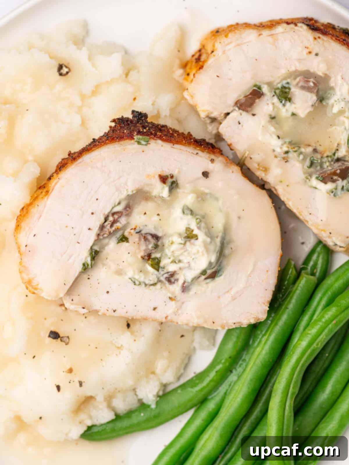 Savory Stuffed Turkey Roll 12 Two enticing slices of stuffed turkey roll are elegantly presented on a plate, accompanied by creamy mashed potatoes and vibrant green beans.