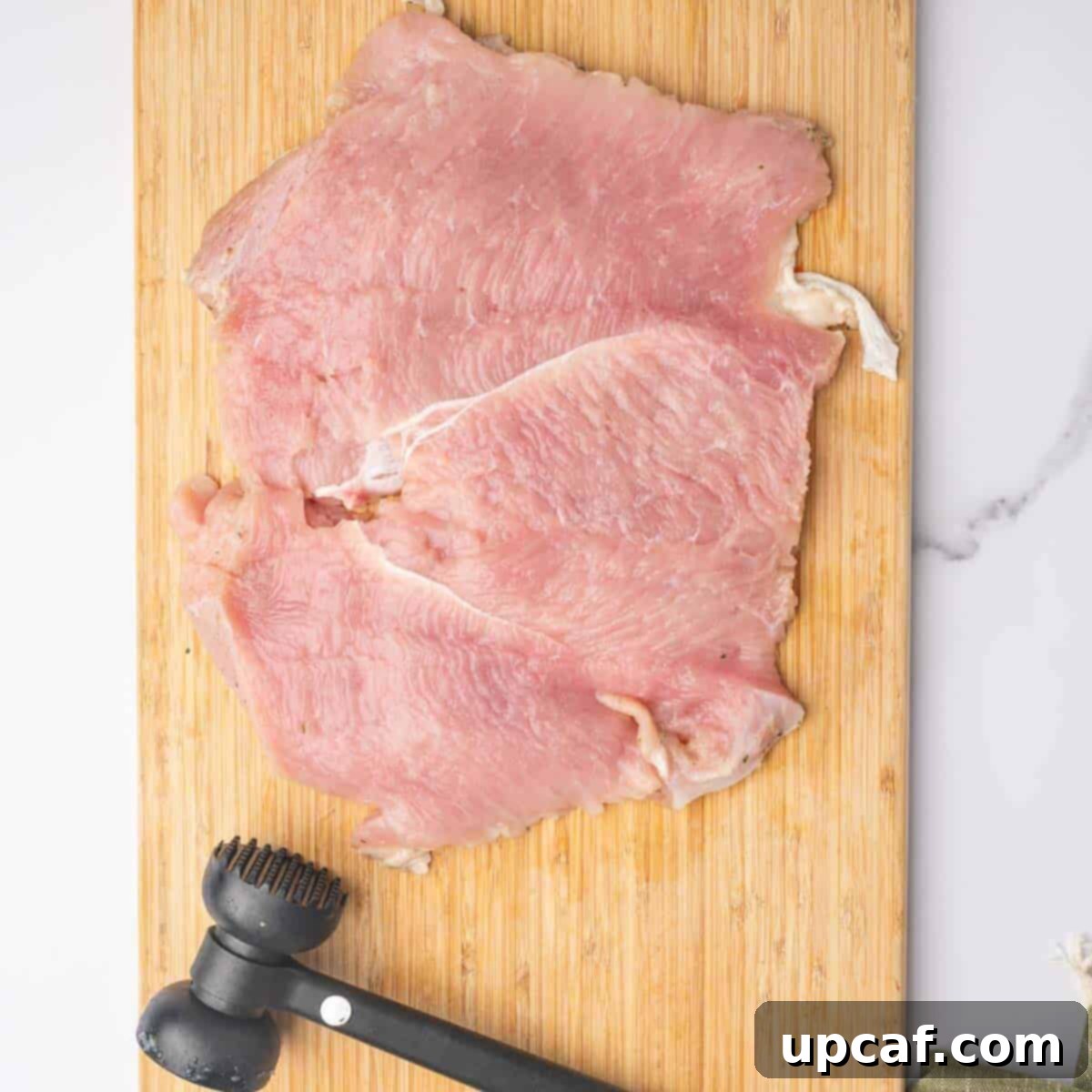 Savory Stuffed Turkey Roll 4 A turkey breast is carefully pounded to an even thickness using a meat mallet on a cutting board, preparing it for stuffing and rolling.