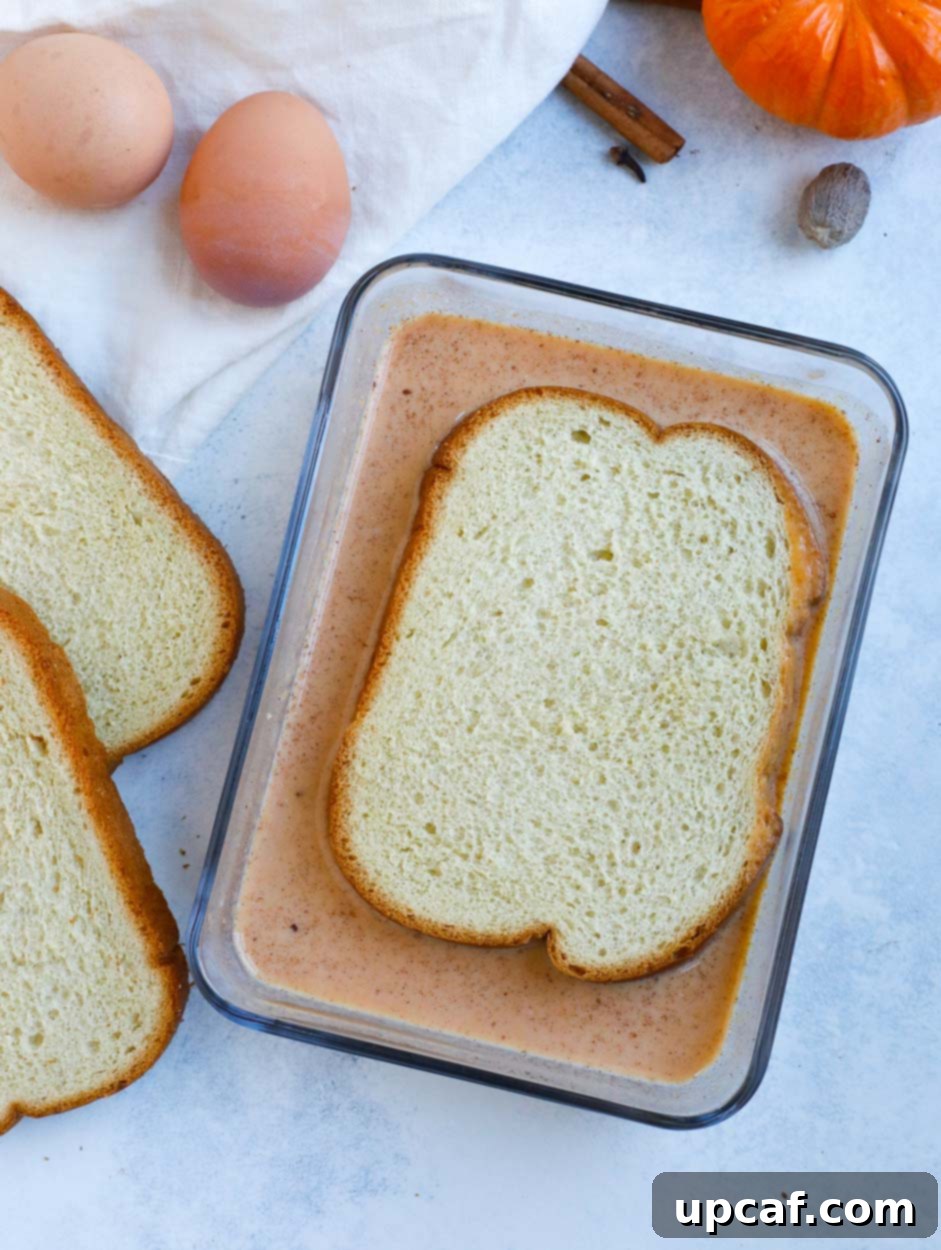 Thick brioche bread slice being dredged in a glass bowl of pumpkin spice French toast batter.