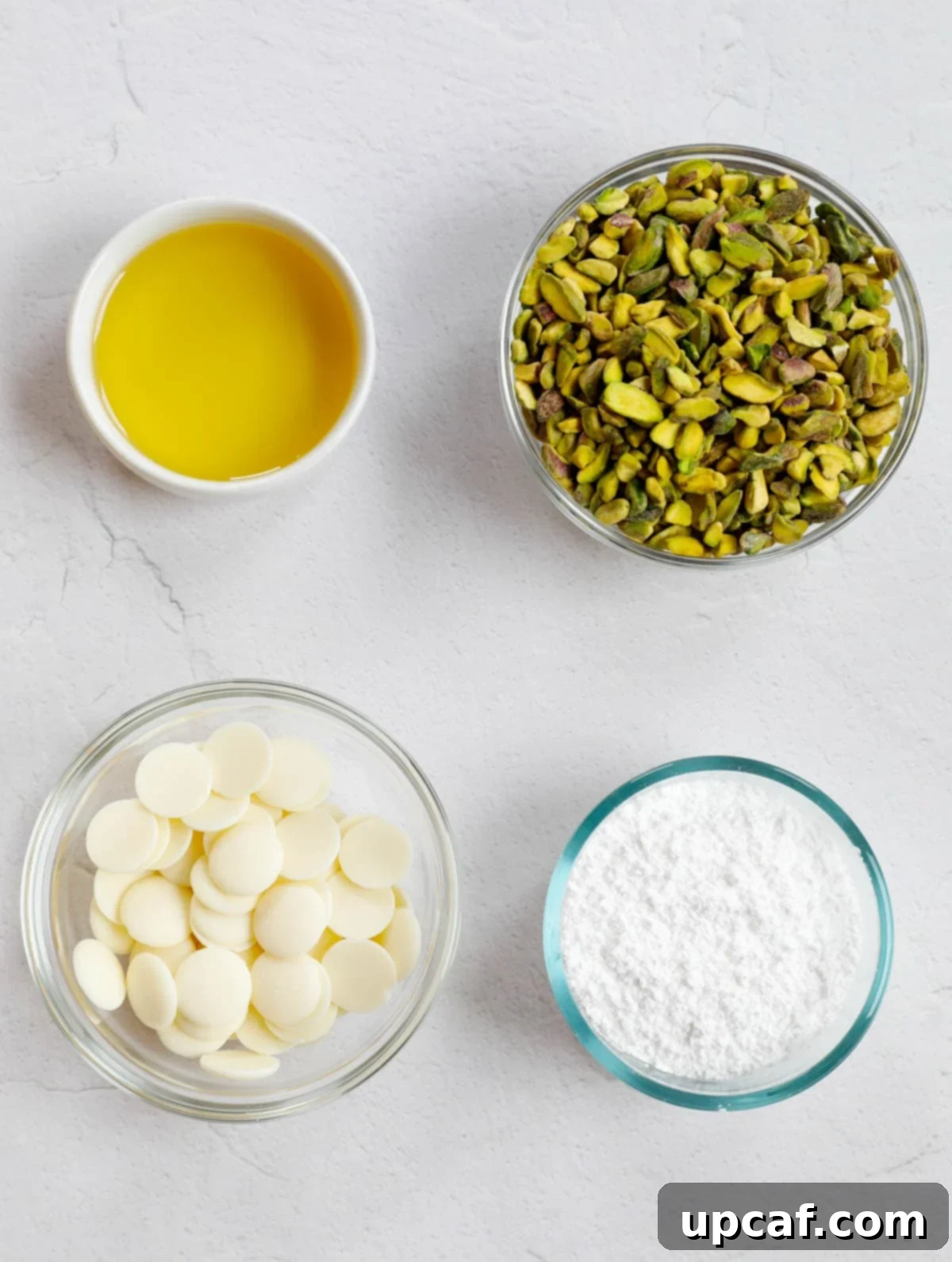 Velvety Pistachio Cream 3 Ingredients needed for pistachio cream: raw pistachios, avocado oil, white chocolate chips, and powdered sugar, laid out on a white surface.