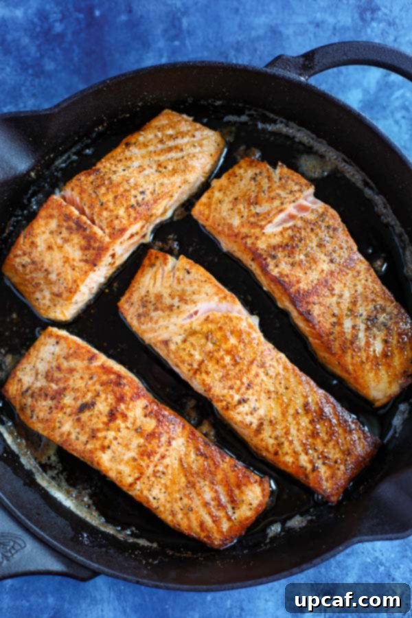 A cast iron skillet containing four salmon fillets expertly cooking on a stovetop, surrounded by a bright blue kitchen counter.