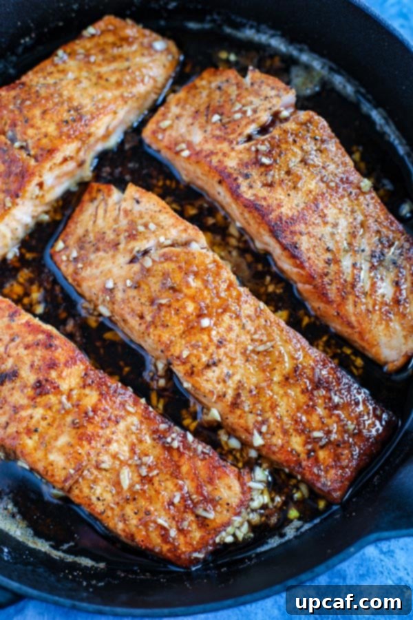 A close-up view of three succulent salmon fillets, coated in a glistening spicy honey garlic sauce, perfectly searing in a cast iron skillet on a blue countertop.