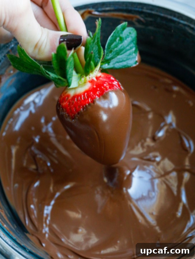 A close-up shot of a hand gently dipping a ripe strawberry into a bowl of rich, melted chocolate, showing the start of its transformation.