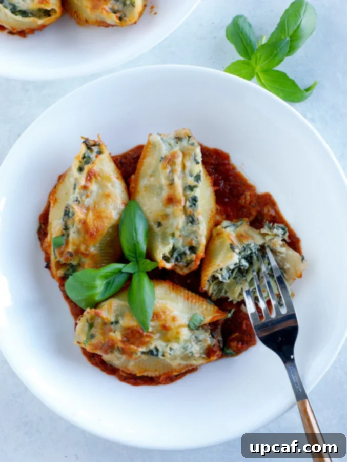 A white bowl with 4 spinach artichoke stuffed shells with a fork and some fresh basil. The shells are glistening with marinara and melted cheese.