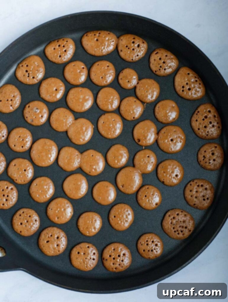Tiny chocolate pancakes cooking on a hot griddle, showing their distinct dark color and small, uniform shape as they bubble and firm up.