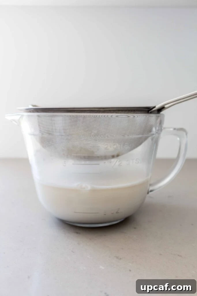 Homemade oat milk being strained through a fine mesh strainer into a bowl, separating the liquid from the oat pulp.